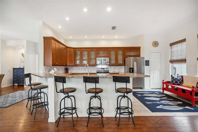 kitchen featuring brown cabinetry, glass insert cabinets, a breakfast bar area, appliances with stainless steel finishes, and light stone counters