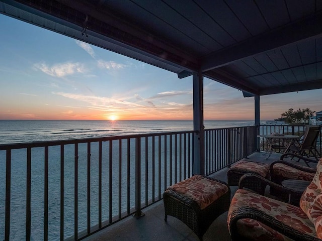 balcony at dusk with a water view, a sunroom, and a view of the beach