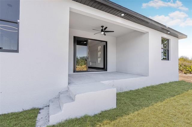 view of exterior entry with a ceiling fan, a patio area, a yard, and stucco siding