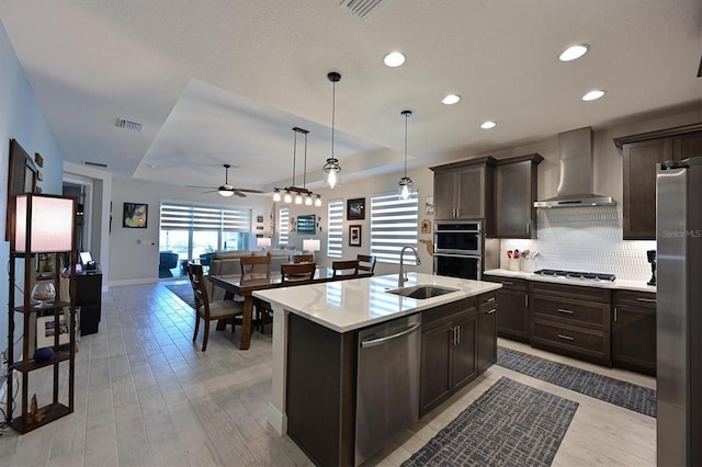 kitchen with a kitchen island with sink, stainless steel appliances, a sink, light countertops, and wall chimney range hood