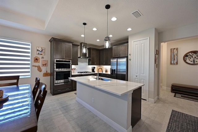 kitchen featuring a kitchen island with sink, stainless steel appliances, dark brown cabinets, wall chimney range hood, and a sink