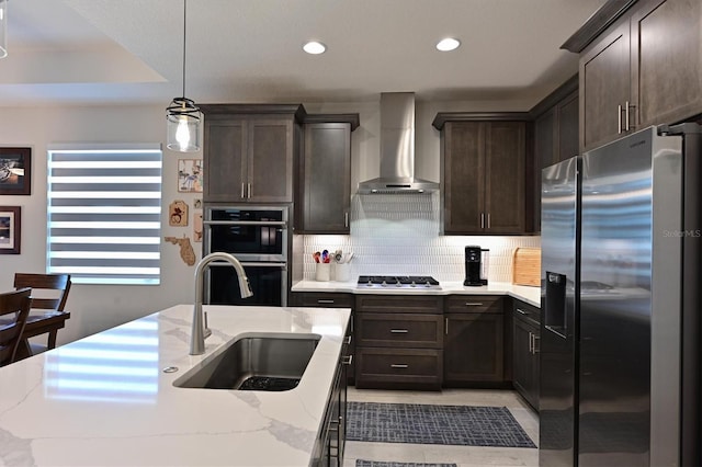 kitchen with wall chimney exhaust hood, appliances with stainless steel finishes, decorative light fixtures, a kitchen island with sink, and a sink