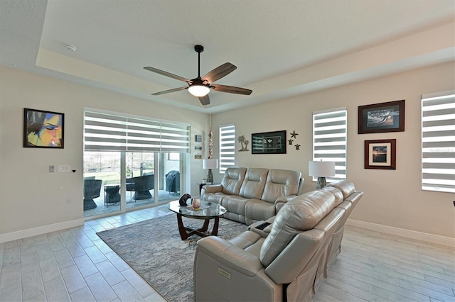 living room featuring a tray ceiling, ceiling fan, light wood-style flooring, and baseboards