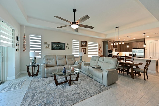 living area with a ceiling fan, light wood-type flooring, a raised ceiling, and baseboards