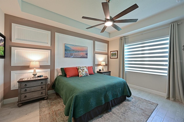 bedroom featuring light wood-type flooring, baseboards, a raised ceiling, and a ceiling fan