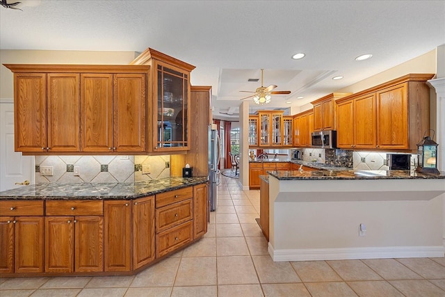 kitchen featuring brown cabinets, stainless steel appliances, glass insert cabinets, dark stone countertops, and a peninsula