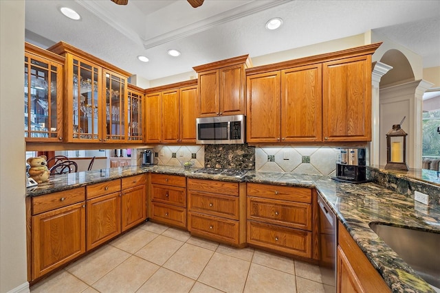 kitchen featuring stainless steel appliances, brown cabinets, dark stone counters, tasteful backsplash, and glass insert cabinets