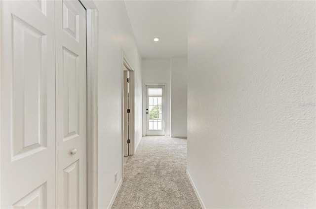 hallway with light carpet, a textured wall, and baseboards