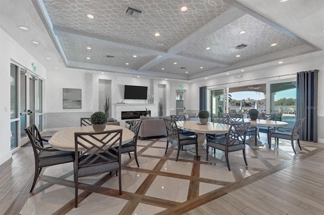 dining area featuring light wood finished floors, visible vents, coffered ceiling, and a glass covered fireplace