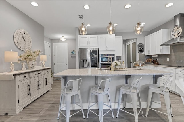 kitchen featuring black electric stovetop, a kitchen island with sink, white cabinets, stainless steel fridge, and wall chimney exhaust hood