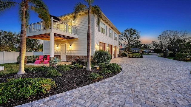 view of property exterior with a balcony, stone siding, an attached garage, fence, and decorative driveway