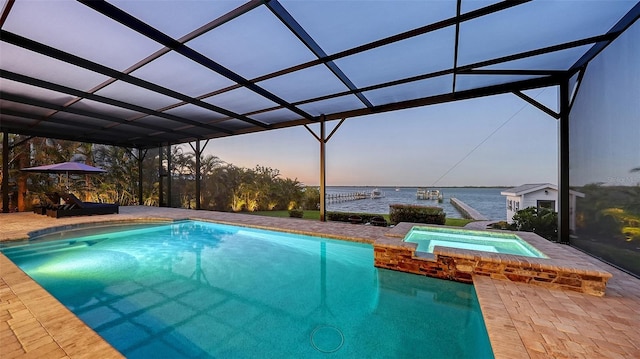 pool at dusk featuring glass enclosure, a patio area, a pool with connected hot tub, and a water view