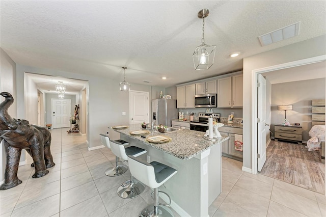 kitchen with an island with sink, light stone counters, hanging light fixtures, stainless steel appliances, and gray cabinetry