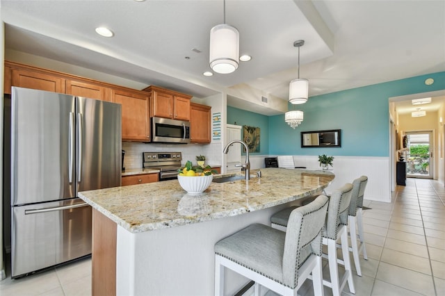 kitchen featuring brown cabinetry, a wainscoted wall, appliances with stainless steel finishes, a sink, and light tile patterned flooring