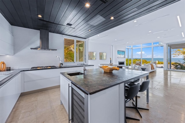 kitchen with white cabinets, an island with sink, wine cooler, modern cabinets, and wall chimney range hood