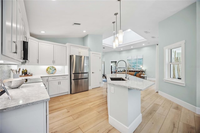 kitchen with a center island with sink, freestanding refrigerator, white cabinetry, a sink, and light stone countertops