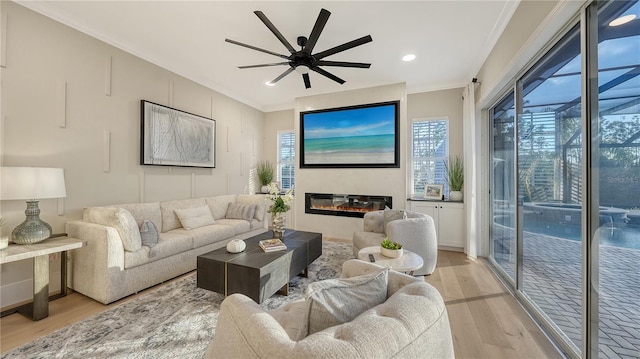 living room with light wood-style floors, a glass covered fireplace, ornamental molding, and a ceiling fan