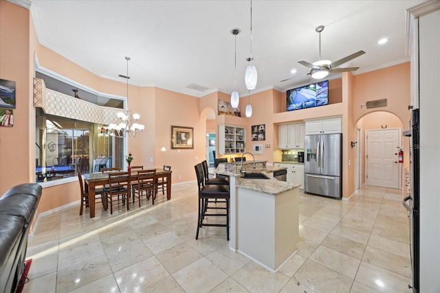 kitchen with stainless steel fridge, arched walkways, light stone counters, ornamental molding, and ceiling fan with notable chandelier