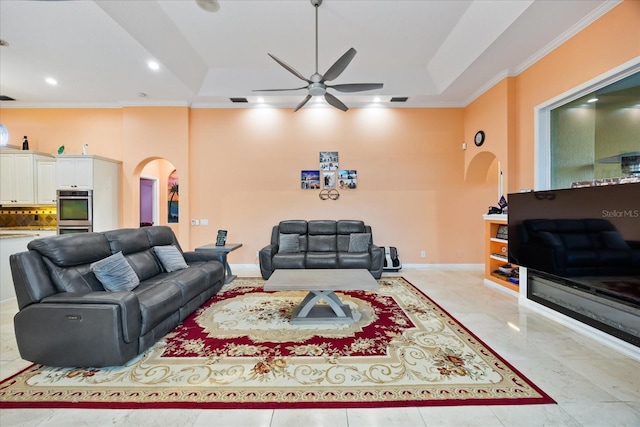 living area with arched walkways, ceiling fan, baseboards, ornamental molding, and a tray ceiling