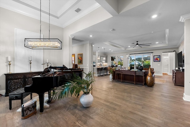 living area with ceiling fan, wood finished floors, visible vents, ornamental molding, and a raised ceiling