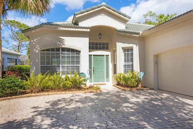 view of exterior entry with a garage, a tile roof, decorative driveway, and stucco siding