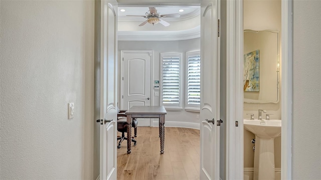 corridor featuring a textured wall, light wood-style flooring, baseboards, ornamental molding, and a tray ceiling