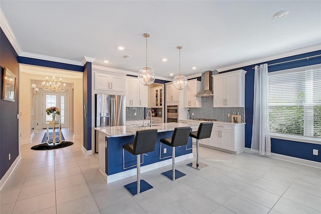 kitchen featuring wall chimney exhaust hood, appliances with stainless steel finishes, a kitchen island with sink, and decorative light fixtures