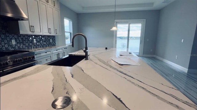 kitchen with a sink, a healthy amount of sunlight, wall chimney range hood, decorative backsplash, and a tray ceiling