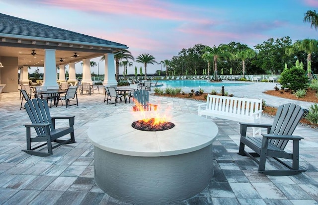 patio terrace at dusk with an outdoor fire pit, a community pool, and a ceiling fan