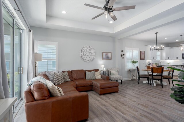 living room featuring ceiling fan with notable chandelier, baseboards, a raised ceiling, and light wood-style floors