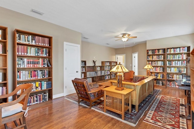 living area with visible vents, ceiling fan, wall of books, and wood finished floors