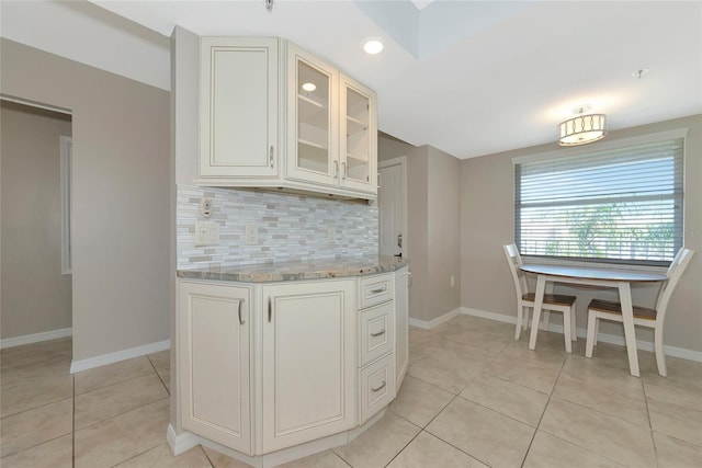 kitchen with tasteful backsplash, baseboards, glass insert cabinets, light stone countertops, and light tile patterned flooring