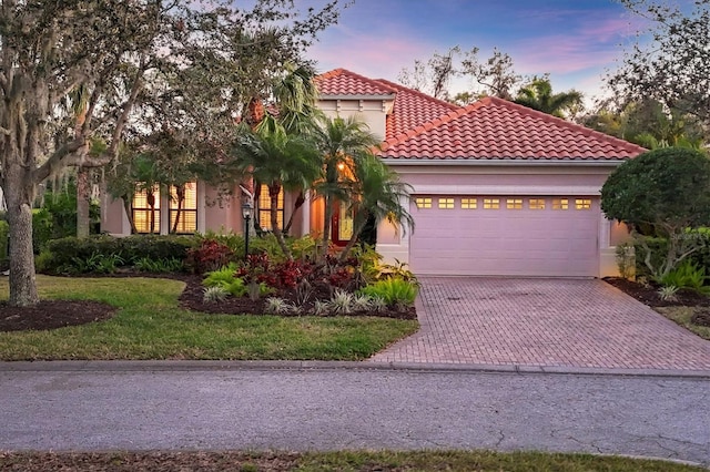 mediterranean / spanish-style home with an attached garage, a tiled roof, decorative driveway, stucco siding, and a front lawn