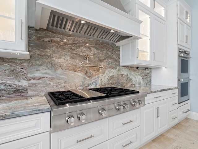 kitchen with wall chimney exhaust hood, light stone countertops, glass insert cabinets, and stainless steel appliances