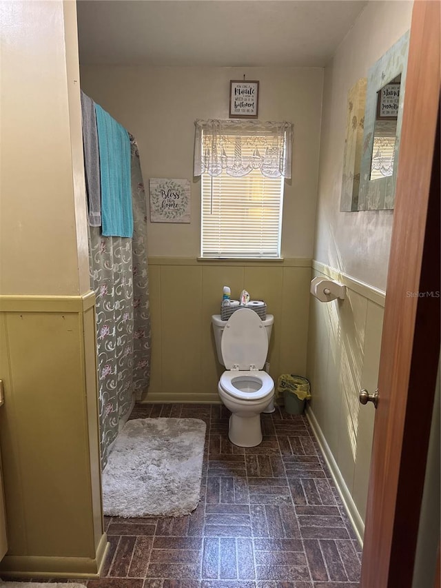 full bath with curtained shower, toilet, brick floor, and wainscoting