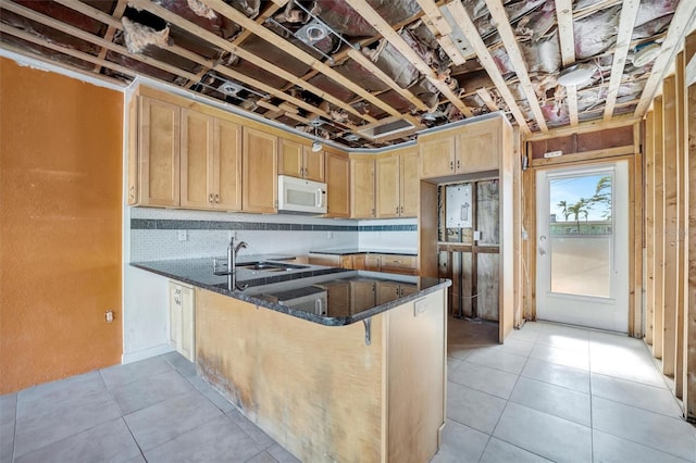 kitchen with light brown cabinetry, white microwave, a sink, dark stone countertops, and a peninsula