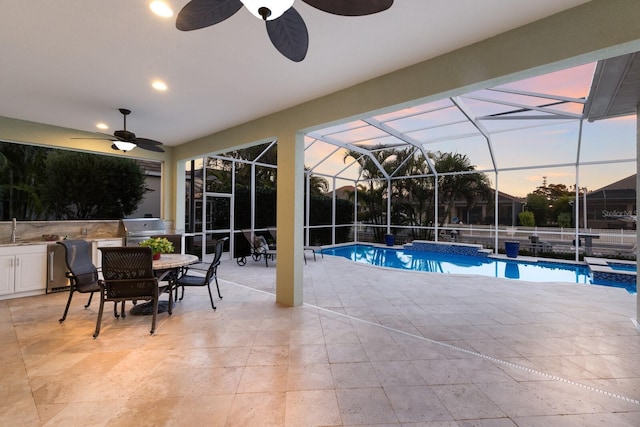 pool at dusk featuring an outdoor kitchen, a ceiling fan, a patio, a lanai, and a sink