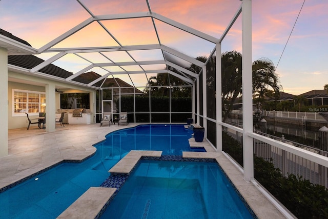 pool at dusk featuring a pool with connected hot tub, a patio, an outdoor kitchen, and a lanai