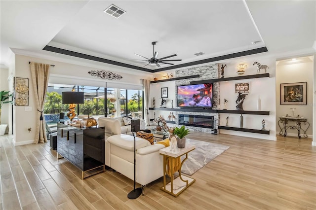 living area featuring light wood finished floors, a raised ceiling, visible vents, a ceiling fan, and a glass covered fireplace