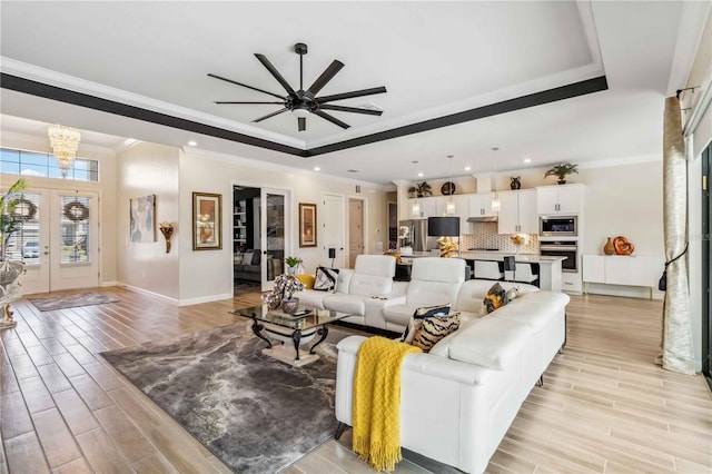 living room featuring a tray ceiling, light wood-style floors, ornamental molding, baseboards, and ceiling fan with notable chandelier