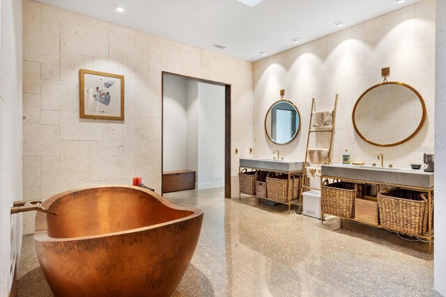 full bath featuring a freestanding bath, speckled floor, two vanities, and tile walls