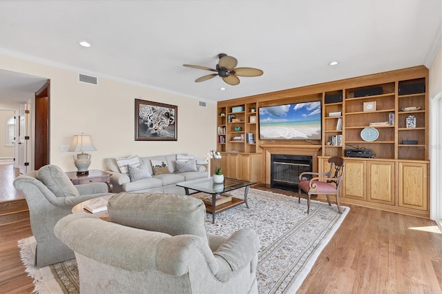 living room with crown molding, light wood finished floors, visible vents, a ceiling fan, and a glass covered fireplace