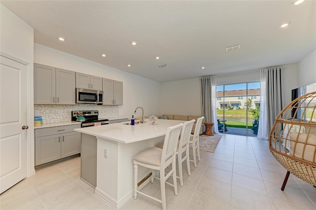 kitchen featuring light countertops, decorative backsplash, gray cabinetry, appliances with stainless steel finishes, and an island with sink