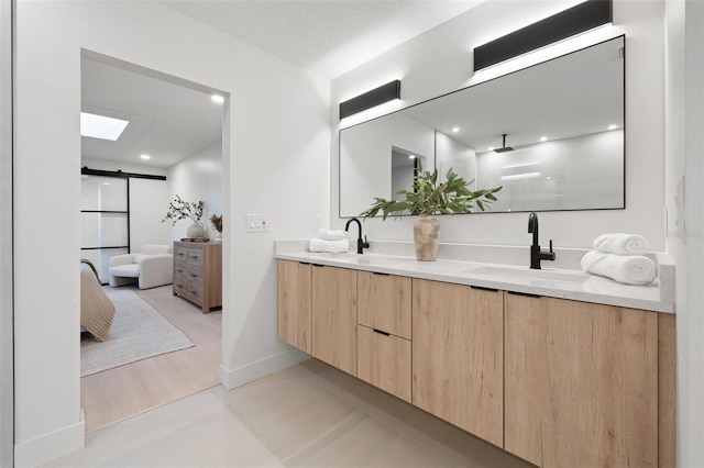 ensuite bathroom featuring a skylight, double vanity, connected bathroom, a sink, and baseboards