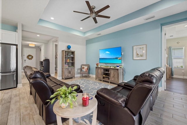 living area with ceiling fan, visible vents, baseboards, a tray ceiling, and light wood finished floors