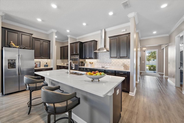 kitchen with appliances with stainless steel finishes, light wood-style floors, a sink, an island with sink, and wall chimney exhaust hood