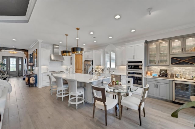 kitchen featuring stainless steel appliances, light countertops, a kitchen island with sink, wall chimney range hood, and beverage cooler