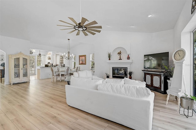 living area with light wood finished floors, a glass covered fireplace, vaulted ceiling, and ceiling fan with notable chandelier