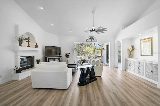 living area featuring light wood finished floors, ceiling fan, baseboards, a glass covered fireplace, and high vaulted ceiling