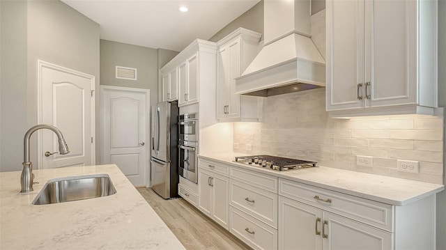 kitchen featuring light wood finished floors, white cabinets, custom range hood, appliances with stainless steel finishes, and a sink